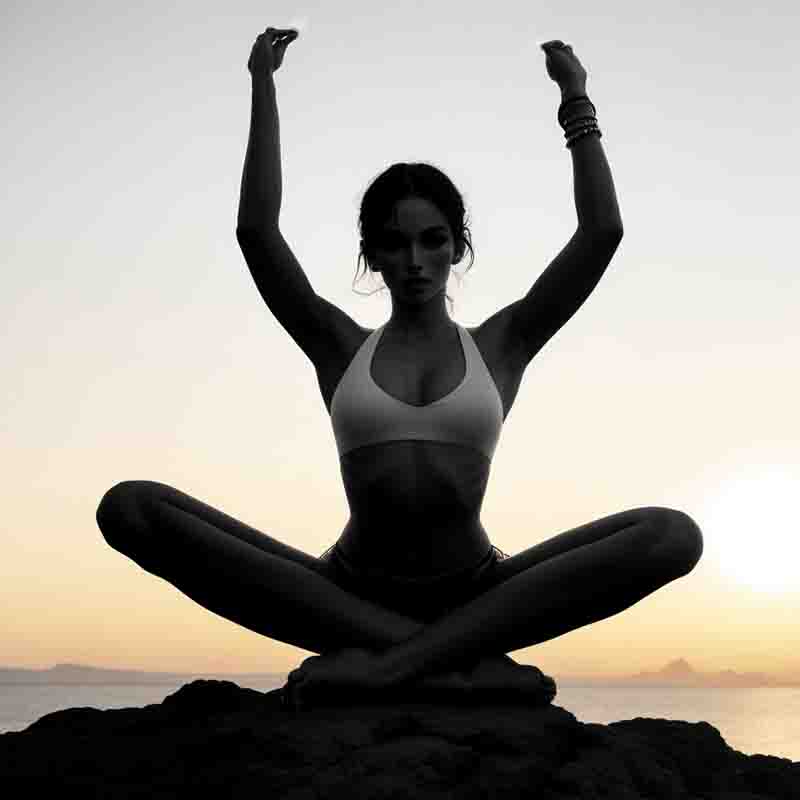 A tranquil scene of a sensual woman in a yoga pose, seated on a rock in a cross-legged position, with their hands resting gently on their knees, silhouetted against the backdrop of a breathtaking Ibiza sunrise at the sea.