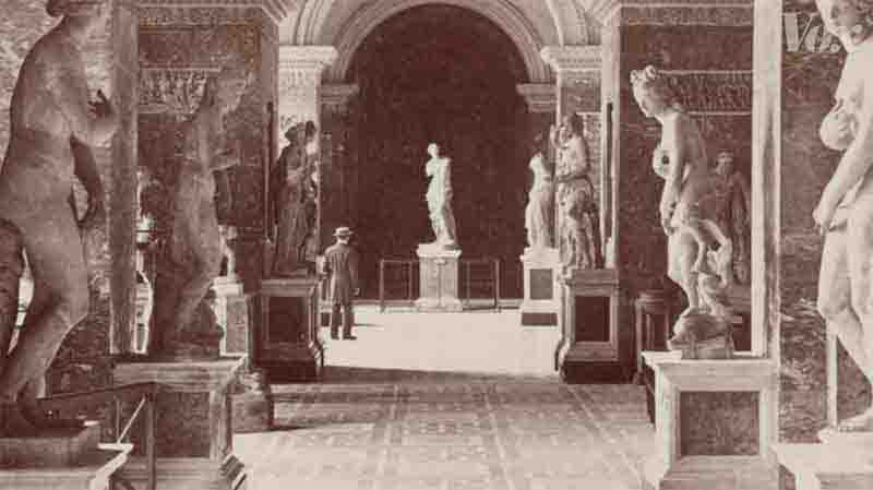 Photo of a man standing in a room filled with statues. The room has a high ceiling and is decorated with arches and columns. The statues are of various sizes and styles, and are arranged along the walls and on pedestals.
