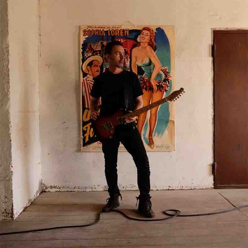 Multiinstrumentalist and Music Producer Likewolf playing an electric guitar with a sunburst finish. Likewolf is standing in a room with a white wall that has a colorful poster featuring “Sophia Loren”. He is dressed in a black t-shirt, black pants, and black shoes. A black cable runs from the guitar to an amplifier, set on a wooden floor.