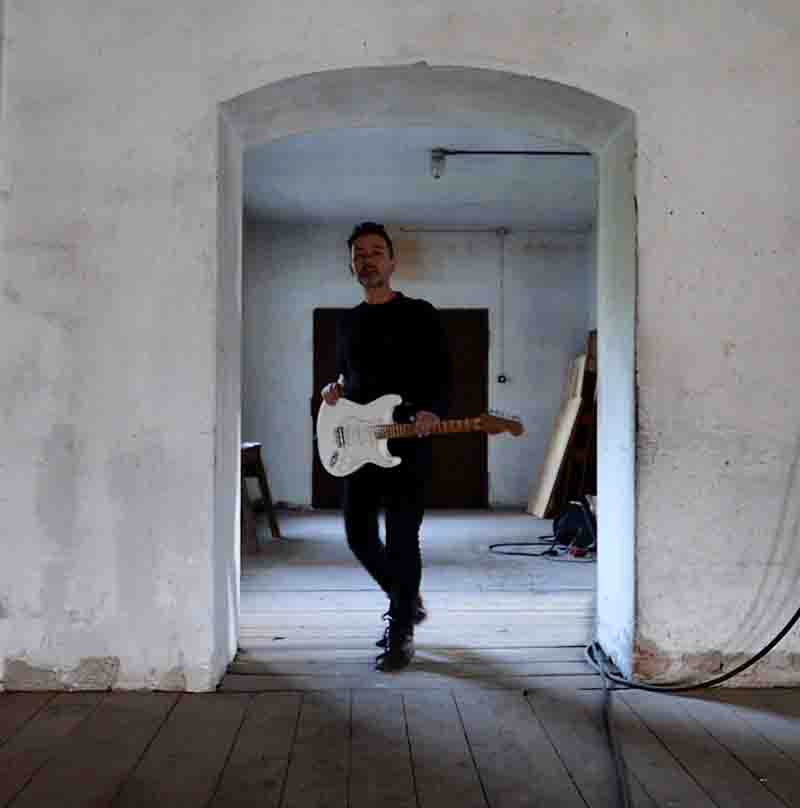 Multiinstrumentalist and Music Producer Likewolf standing in an archway, holding a white electric guitar. Likewolf is dressed in a black long sleeve shirt and black pants. A wooden floor underfoot has a cable running across it, hinting at the musical setting.