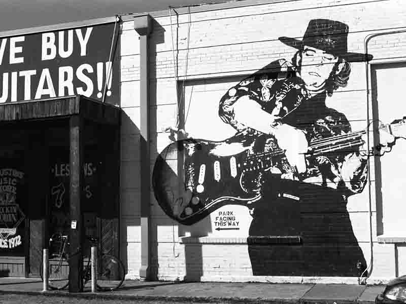 A monochrome image of a guitar store with murial of Stevie Ray Vaughan playing a Fender Stratocaster guitar displayed on the wall