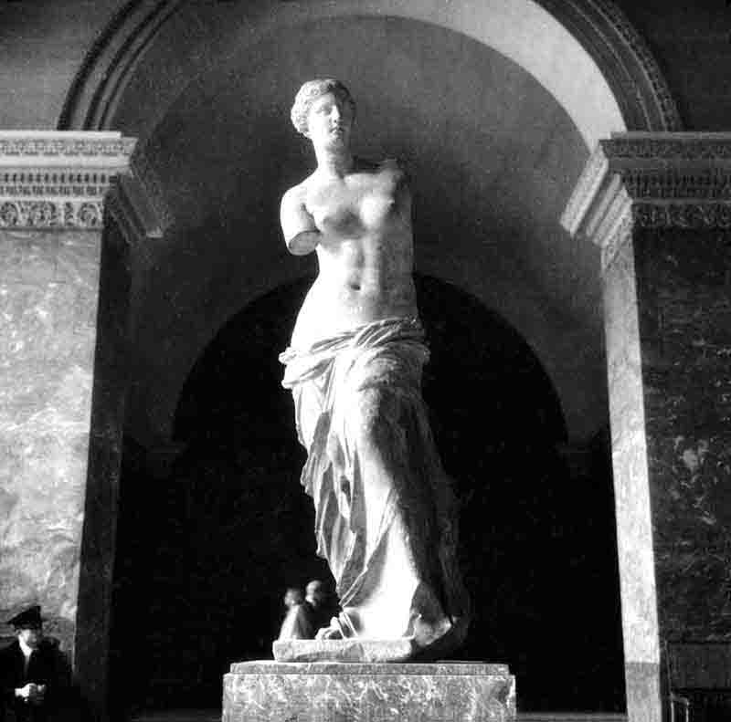 The Venus de Milo with a cloth draped over her lower body. The background is guard of the museum in a room with marble walls and a high ceiling.